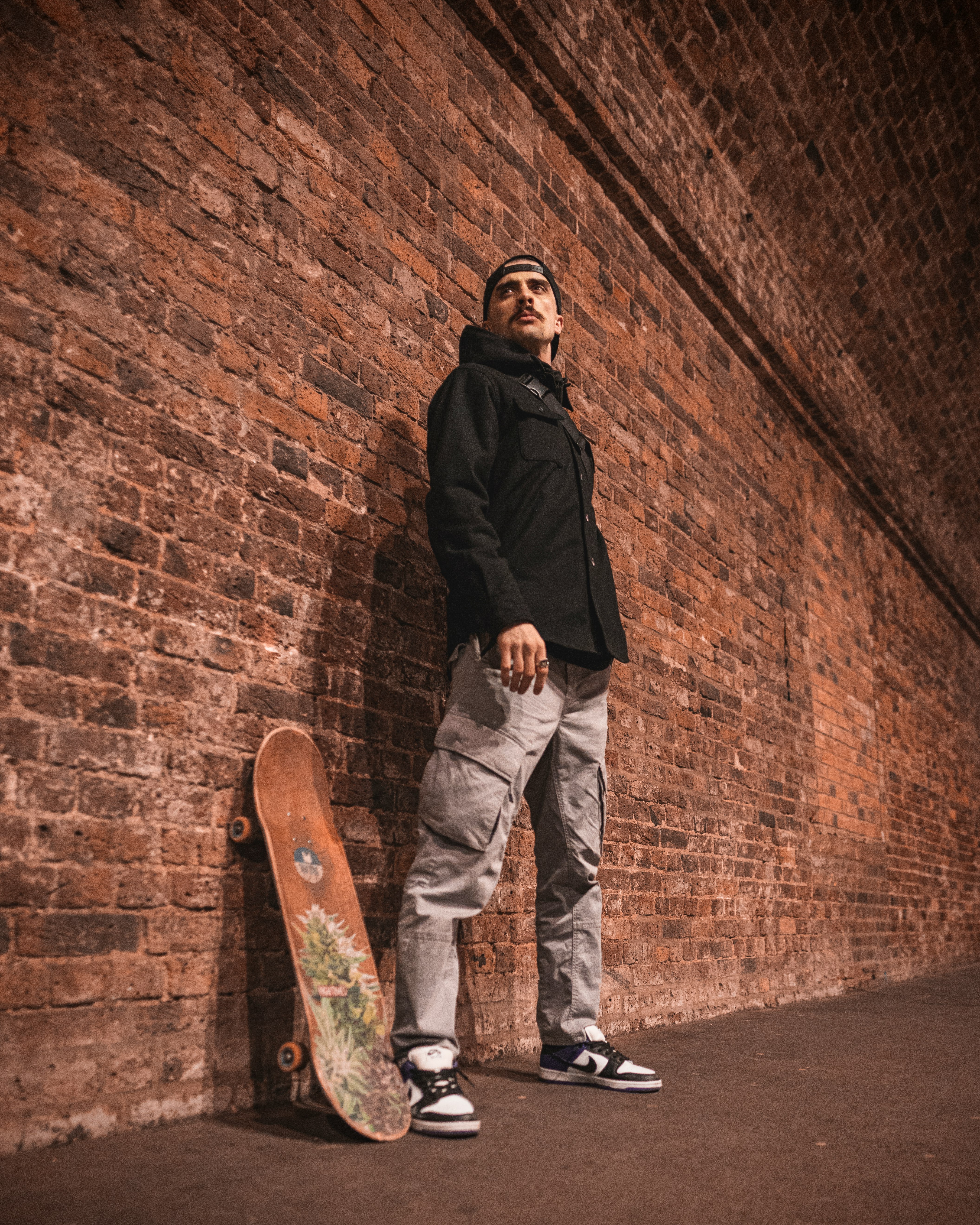 man in black jacket and white pants standing beside brown brick wall